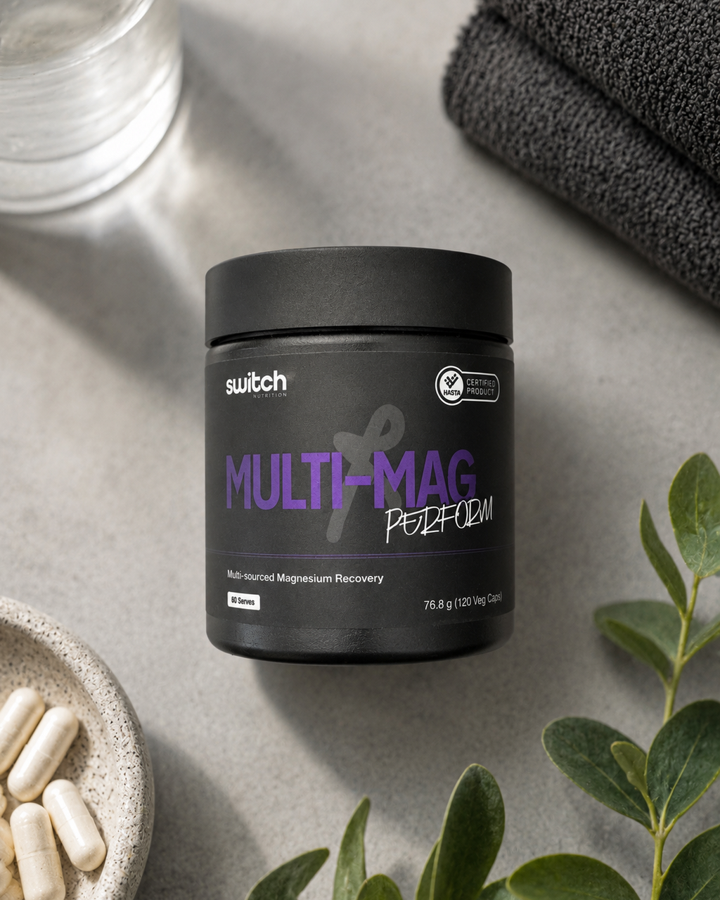 A black jar of SWITCH Multi-Mag Potassium dietary supplement, styled on a gray surface with folded dark towels, green leaves, and capsules in a bowl.