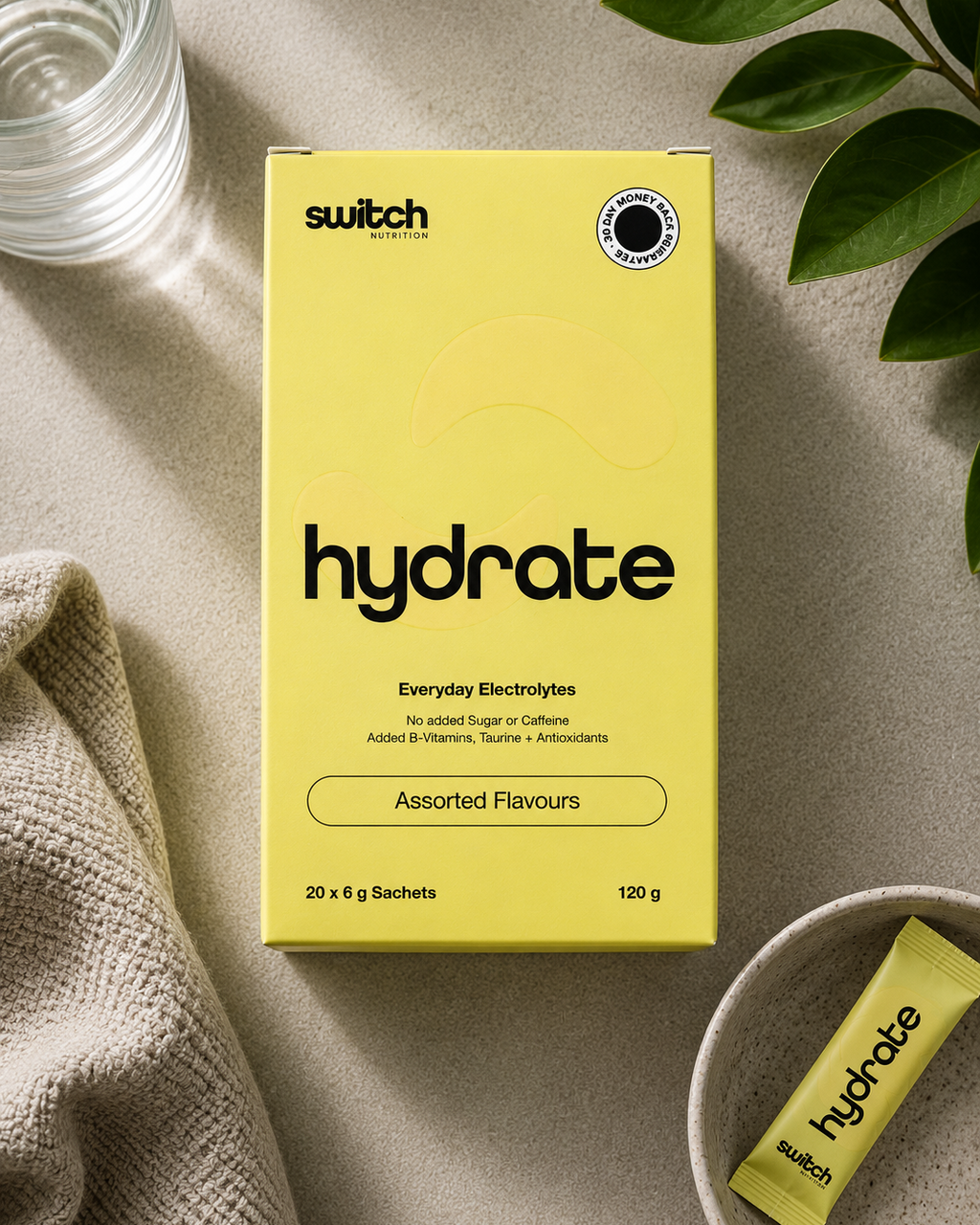 A yellow box of Switch Hydrate Everyday Electrolytes, shown in a styled flat-lay with a glass of water, a beige cloth, and green leaves on a neutral textured surface.