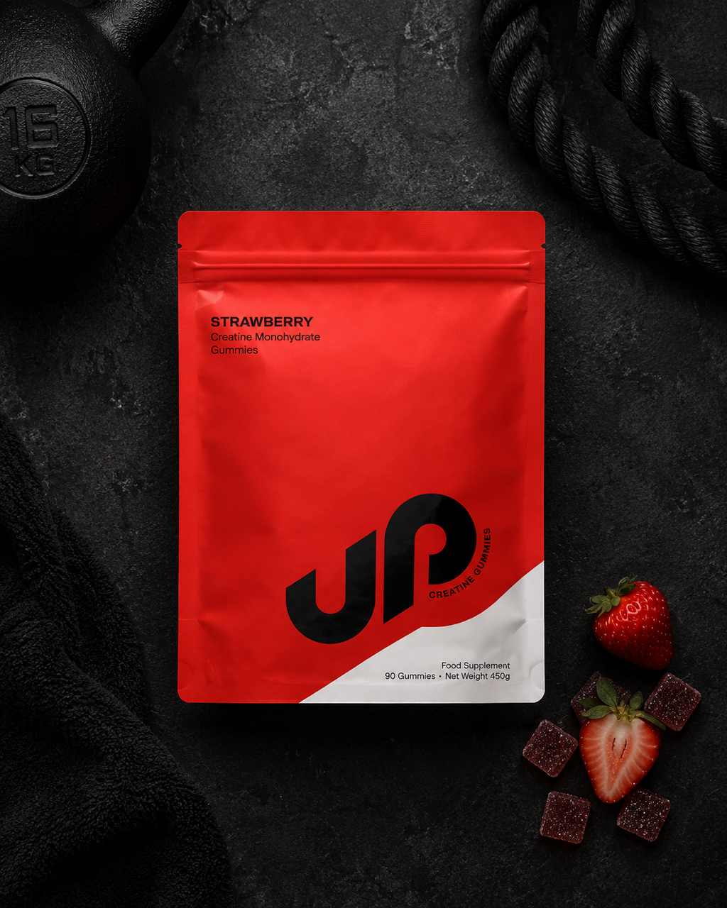 A red stand-up pouch of UP Nutrition Strawberry Creatine Microdose Gummies, styled on a dark textured background with a dumbbell, rope, and fresh strawberries and gummy candies.