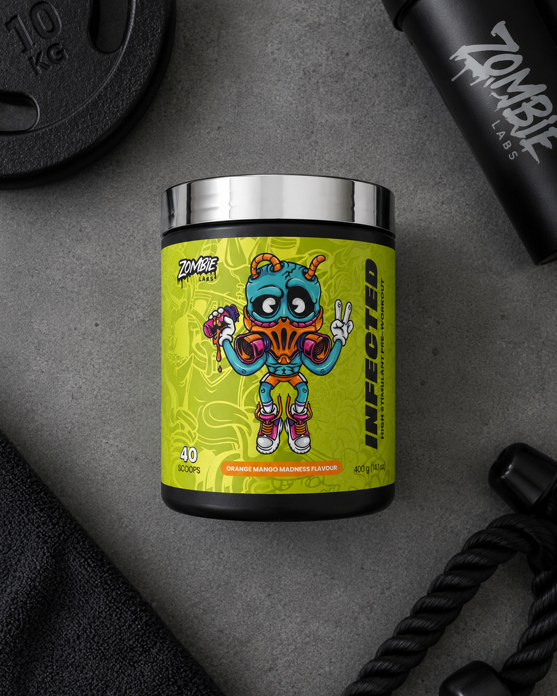 A black supplement tub with a bright yellow label featuring a colorful cartoon zombie character, styled on a gray surface with gym props including a weight plate, bottle, and rope.