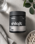 A black jar of switch brand Shilajit supplement placed on a light gray stone surface, surrounded by a glass of water, green leaves, a textured cloth, and a bowl of capsules.