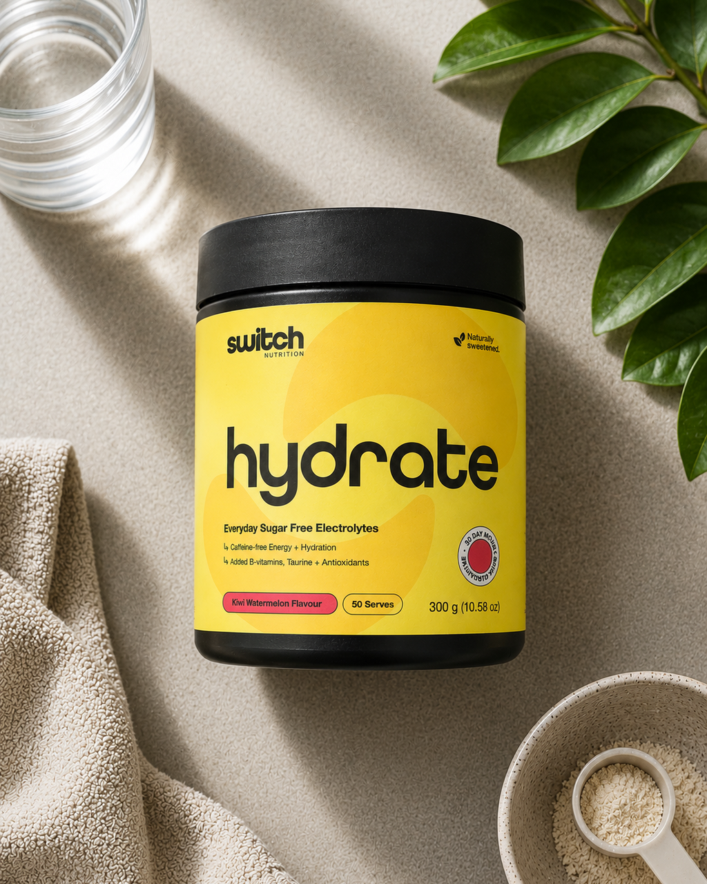 A tub of switch switch hydrate sugar-free electrolytes powder, styled in a flat-lay with a glass of water, a beige towel, a bowl of powder, and green leaves on a textured beige surface.